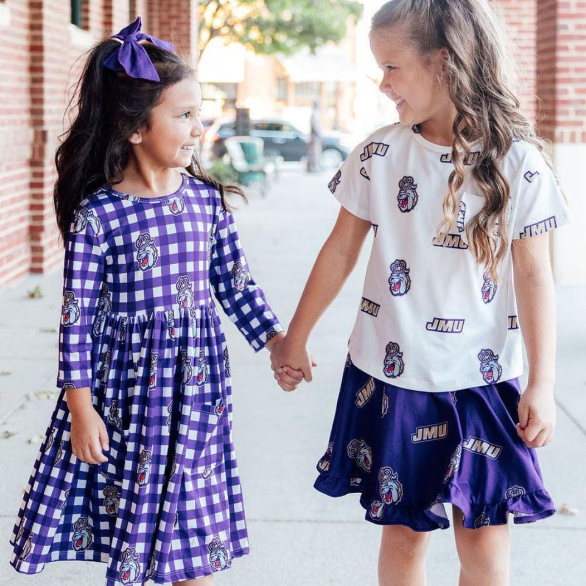 Two children holding hands, one in a purple checkered dress and the other in a white t-shirt and navy shorts with JMU logos.