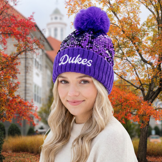 Woman wearing a purple 'Dukes' beanie with a purple pom-pom in an autumn setting.