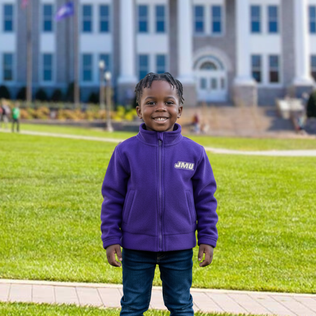 Child wearing a purple jacket with 'JMU' on it, standing on grass in front of a building.