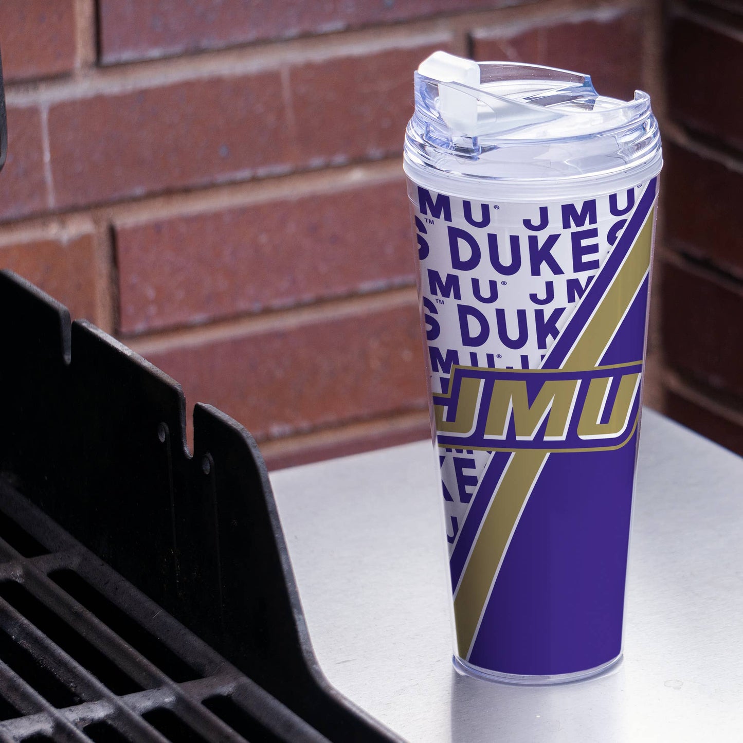 Tumbler with 'JMU DUKES' design on a surface with a brick wall background