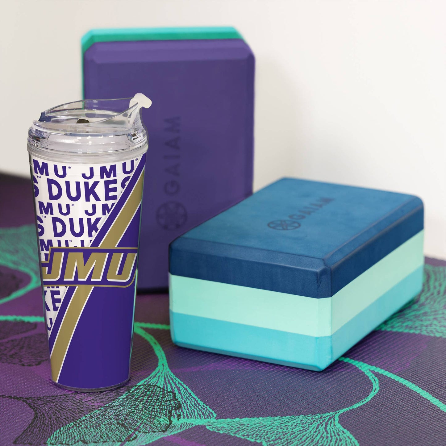 Tumbler with 'JMU' design, blue and green yoga block, and purple block on a mat.