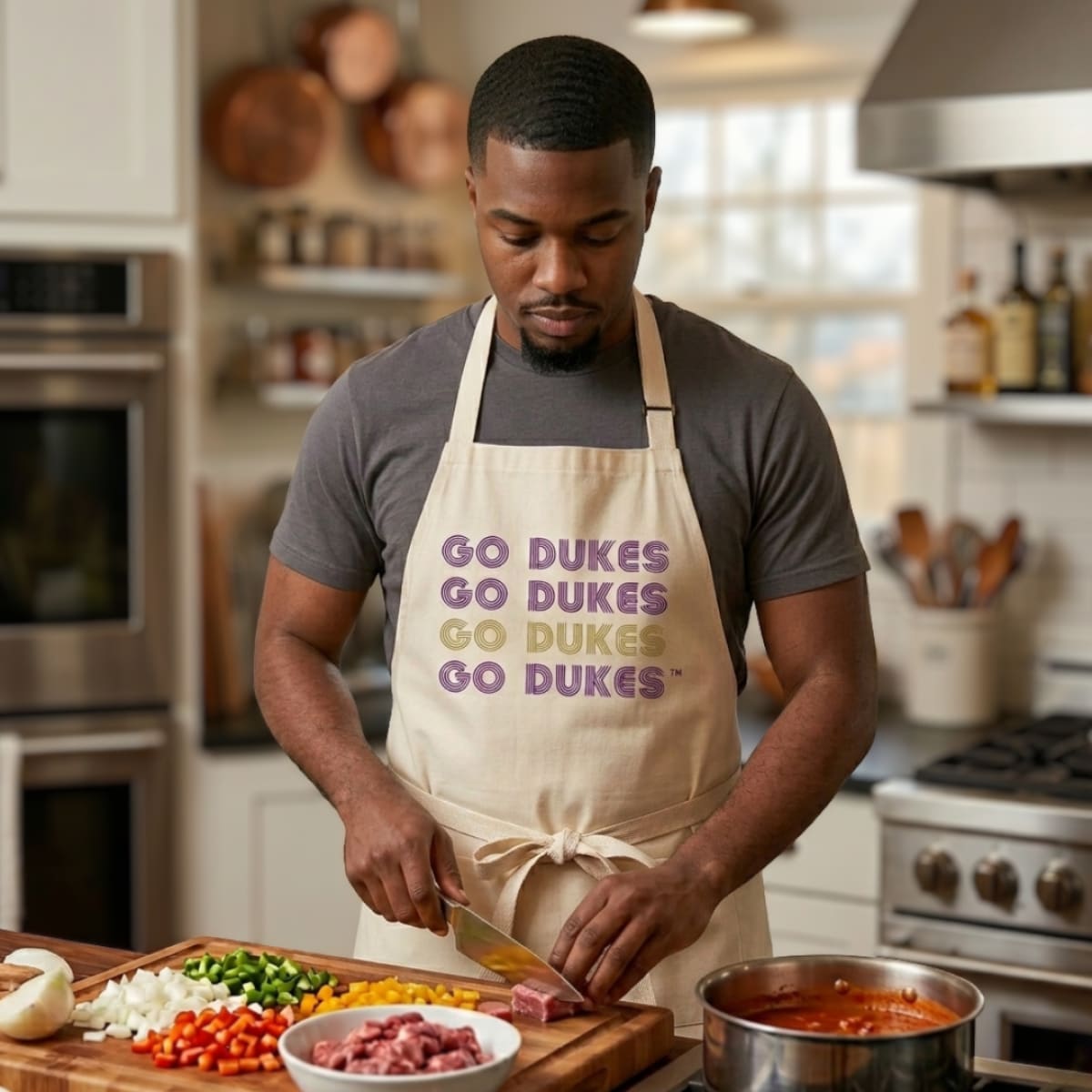 Man in a kitchen wearing a 'Go Dukes' apron, preparing food.