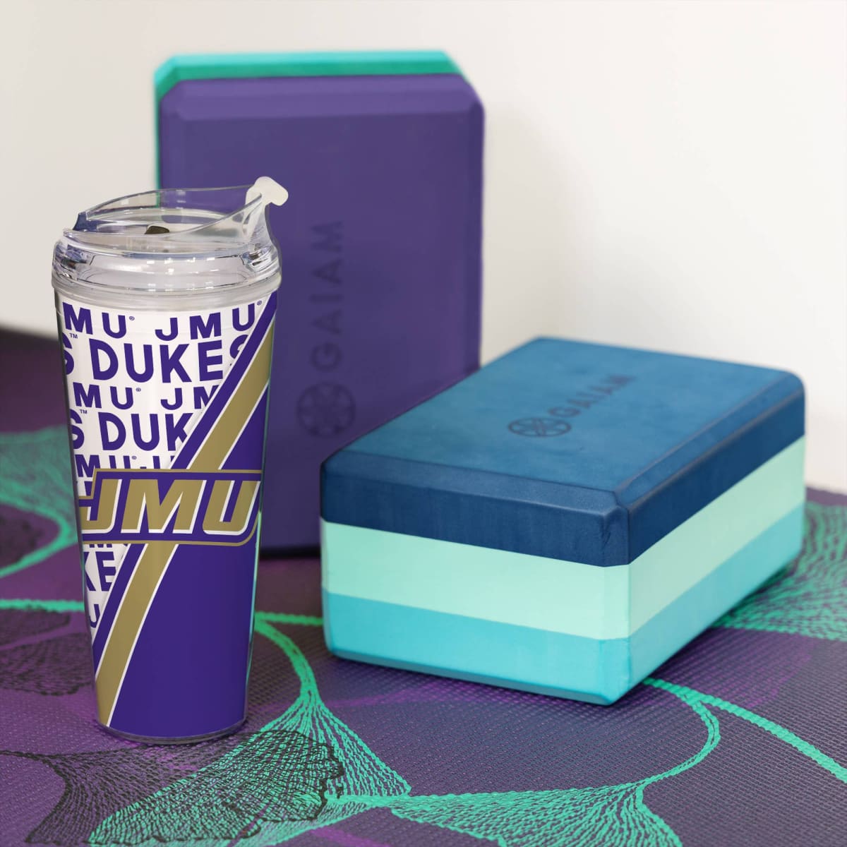 Tumbler with 'JMU' design, blue and green yoga block, and purple block on a mat.