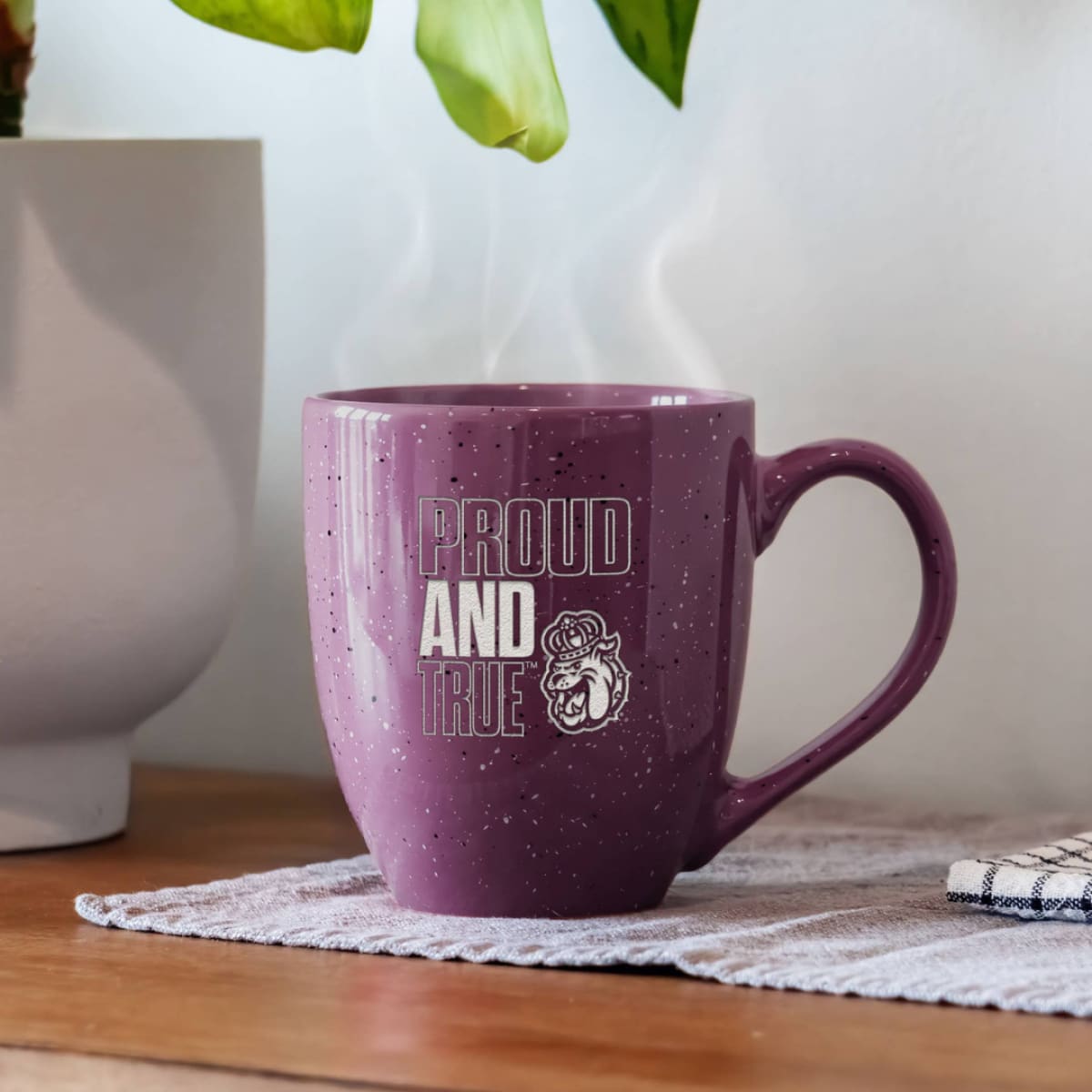 Purple mug with 'Proud and True' text on a wooden surface with a plant in the background