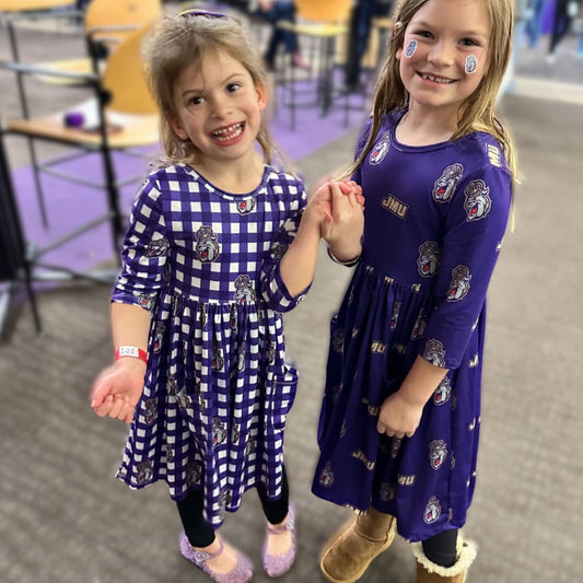 Two young girls in matching blue dresses with logos, holding hands indoors.