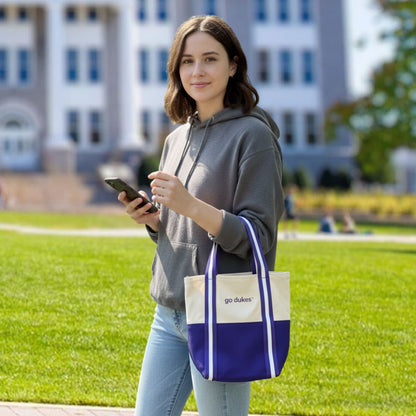 Woman holding a phone and a tote bag with 'go dukes' text on a grassy area.