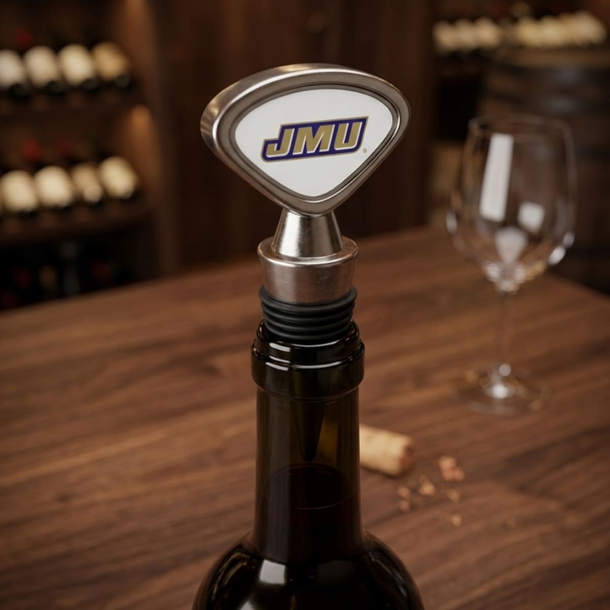 Wine bottle stopper with JMU logo on a wooden table with wine bottles and glasses in the background.